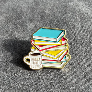 Das Produktbild zeigt einen Bücherstapel-Pin, vor dem ein Kaffeebecher mit der Aufschrift "sorry, i'm booked" steht. Die Farben der Bücher sind Hellblau, Rot und Gelb. Die Tasse ist weiß. Die Umrandung und die Schrift auf der Tasse sind golden.